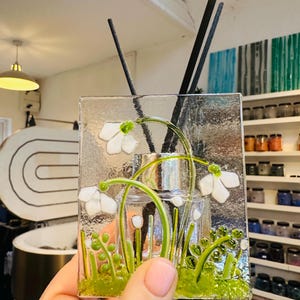 May include: A clear glass diffuser with a floral design featuring white flowers and green stems. The diffuser has a silver cap and includes black diffuser sticks. The design is set against a textured glass background.