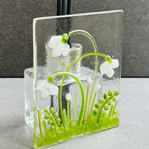 May include: A clear glass art piece with a floral design. White flowers with green stems and leaves are set against a clear background. The rectangular piece is likely a decorative item.