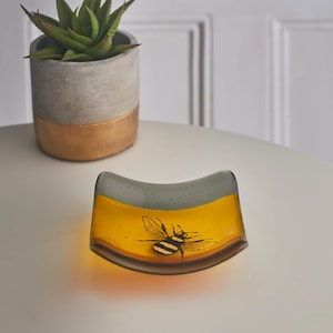 May include: A small, curved glass dish with a yellow and gray design. A black and yellow bee is painted on the yellow portion of the dish.