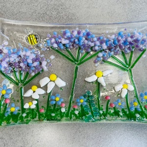 May include: A glass dish with a floral design featuring purple and white flowers, green stems, and a small yellow bee.