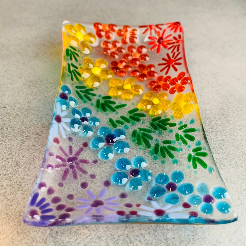 Puede incluir: Un plato de vidrio transparente con un dise&ntilde;o floral de arco&iacute;ris. El plato est&aacute; decorado con peque&ntilde;as flores y hojas de vidrio de colores.