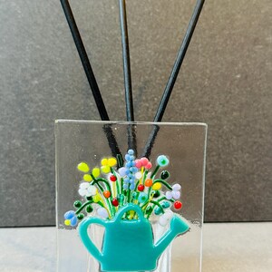 May include: A glass diffuser with a teal watering can design and colourful flowers. Black diffuser sticks are in the glass.