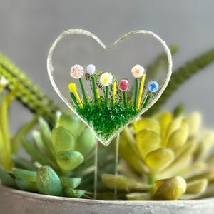 May include: A clear glass heart-shaped plant stake with a miniature garden design. The garden features colorful flowers and green grass. The stake is designed to be inserted into a potted plant, adding a decorative touch.