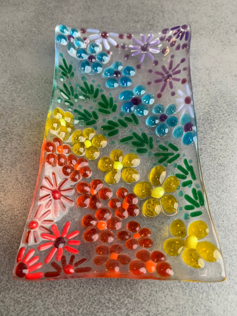 Puede incluir: Un plato de vidrio transparente decorado con peque&ntilde;as flores de vidrio en tonos de rojo, naranja, amarillo, verde, azul y morado.