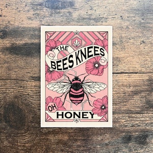 Puede incluir: Una impresión artística rectangular rosa y blanca con una ilustración de abeja. La abeja tiene rayas negras y rosas. La impresión incluye el texto "THE BEES KNEES OH HONEY" con elementos florales y geométricos.