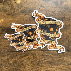 May include: Three black and yellow skull stickers with the text "Without darkness there can be no light" on each. The stickers are on a wooden surface.