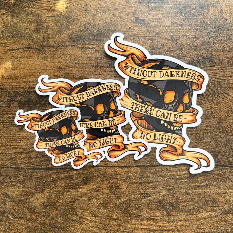 May include: Three black and yellow skull stickers with the text "Without darkness there can be no light" on each. The stickers are on a wooden surface.