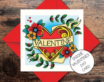 May include: A Valentine's Day card featuring a red heart tattoo design. The heart is surrounded by flowers and a banner that reads "VALENTINE". The card has a blue background and a white border.