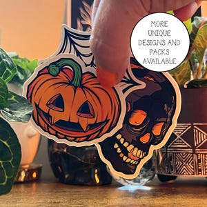 May include: Two Halloween themed stickers, one of a jack-o'-lantern with a toothy grin and the other of a skull with a toothy grin. The stickers are white with black outlines and are on a wooden surface.