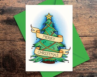 May include: A Christmas card featuring a green Christmas tree with a gold star on top. The tree is decorated with red and gold garland and lights. A banner with the words "MERRY CHRISTMAS" is wrapped around the tree. The card is on a wooden surface with a green envelope.