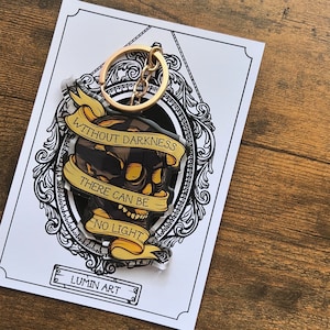 May include: A skull-shaped keychain with a gold-colored chain and ring. The skull is black and yellow, with a banner that reads "WITHOUT DARKNESS THERE CAN BE NO LIGHT." The keychain is on a white card with a decorative black border and the text "LUMIN ART."