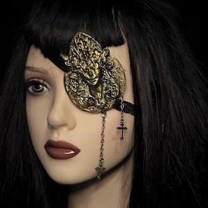 May include: An antique gold eye patch with a detailed face design. A delicate chain with a cross charm hangs from the patch. The eye patch is attached to a black strap, creating a gothic or Victorian aesthetic. The style is reminiscent of the Victorian era.