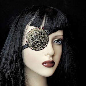 May include: A black eye patch with an ornate gold and red metal design featuring a snake. The design has a raised, textured surface.