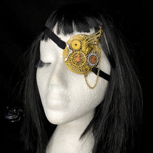 May include: A gold steampunk-style eye patch with gears and a chain. The eye patch is worn on a mannequin with black hair.