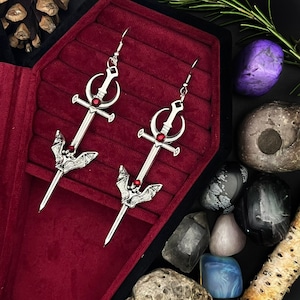 May include: A pair of silver earrings with a sword and bat design. The earrings have red gemstones on the swords. The earrings are displayed in a red velvet jewelry box.