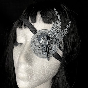 May include: A silver eye patch with a skull and wings design. The eye patch is attached to a black elastic band.
