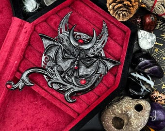 May include: A black metal hair comb with a bat and crescent moon design. The comb has red gemstone accents and is displayed in a red velvet box.