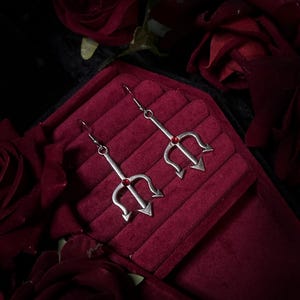 May include: A pair of silver-toned trident earrings with red gemstone accents. The earrings are displayed on a red velvet surface, surrounded by dark red roses. The trident design features three prongs and a small red gem at the center.