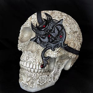May include: A decorative skull with a black bat-shaped mask adorned with red gems. The mask is attached to a black band. The skull is white with floral and leaf patterns.