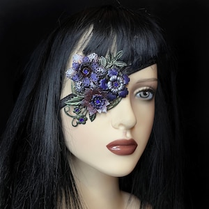 May include: A purple and green floral eye patch with a black velvet strap. The eye patch is decorated with rhinestones and has a delicate, intricate design.
