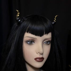 May include: Black headband featuring small, curved horns. The horns are black with gold tips. The headband is worn on a mannequin with black hair and dark lipstick. A unique accessory.