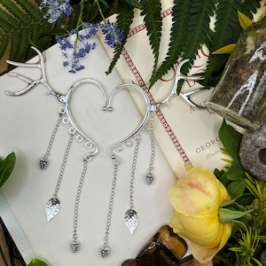 May include: Silver ear cuff with a heart shape and dangling chains with leaf and berry charms. The cuff is on a book with a floral design and the text "GEORGE ILLUSTRATED".