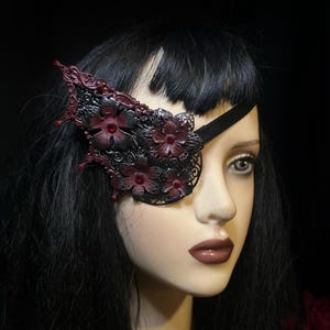 May include: A decorative eye patch featuring a black filigree design with burgundy floral accents and red crystal centers. The eye patch is attached to a black velvet strap. The design is set against a black background.