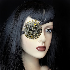 May include: A black eye patch with a gold and black octopus design and yellow rhinestones. The eye patch is made of fabric and has an adjustable strap.