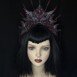 May include: A gothic-style crown in black and red, featuring intricate filigree, crescent moon accents, and a central red gemstone. The crown has wing-like details and dangling chains, set against a black background. A mannequin head with black hair and red lipstick is in the image.