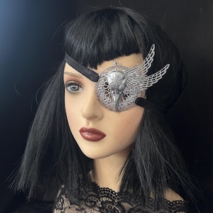 May include: A decorative eye patch featuring a silver-toned bird skull emblem centered on a circular filigree base with an attached wing detail. The piece is secured with a black elastic strap and includes a small clear crystal accent at the top.