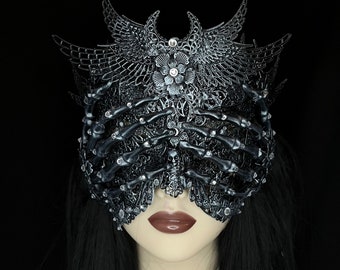 May include: A detailed, dark silver mask with a skeletal hand design covering the eyes. The mask features ornate filigree, a floral centerpiece, and wing-like accents. Rhinestones add sparkle to the gothic-inspired accessory.
