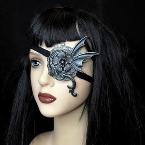 May include: A silver eye patch with a dragon and floral design. The eye patch is worn over a black eye patch strap.