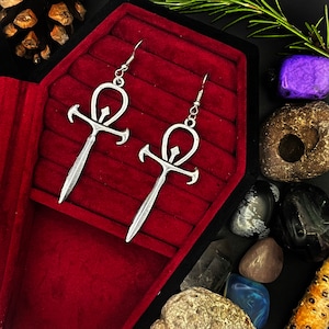 May include: A pair of silver Ankh earrings with a detailed design. The earrings are displayed on a red velvet background.