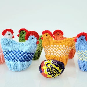 May include: Six knitted chicken egg cosies in various colours, including pink, blue, green, orange, and light blue. The cosies are arranged in a row with a Cadbury Creme Egg in the centre.