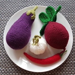 May include: A white plate with four knitted vegetables: a purple aubergine, a red beetroot with green leaves, a white garlic, and a red chilli pepper.