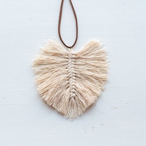 May include: Cream-coloured macrame leaf pendant with a brown cord. The leaf design features a central woven spine with fringed edges, creating a textured, natural aesthetic. The pendant hangs against a white background.