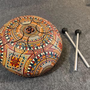 Op de afbeelding: Een handpan drum met een kleurrijk, ingewikkeld mandala-ontwerp in tinten rood, oranje, geel en blauw. Het midden heeft het Om-symbool. Twee grijze drumstokken met zwarte uiteinden zijn inbegrepen.