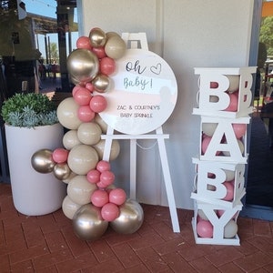 May include: White wooden sign with the text "Oh Baby!" and "Zac & Courtney's Baby Sprinkle".  A white easel holds the sign.  A balloon arch made of pink and beige balloons is on the left.  A white wooden block tower with the letters "BABY" is on the right.