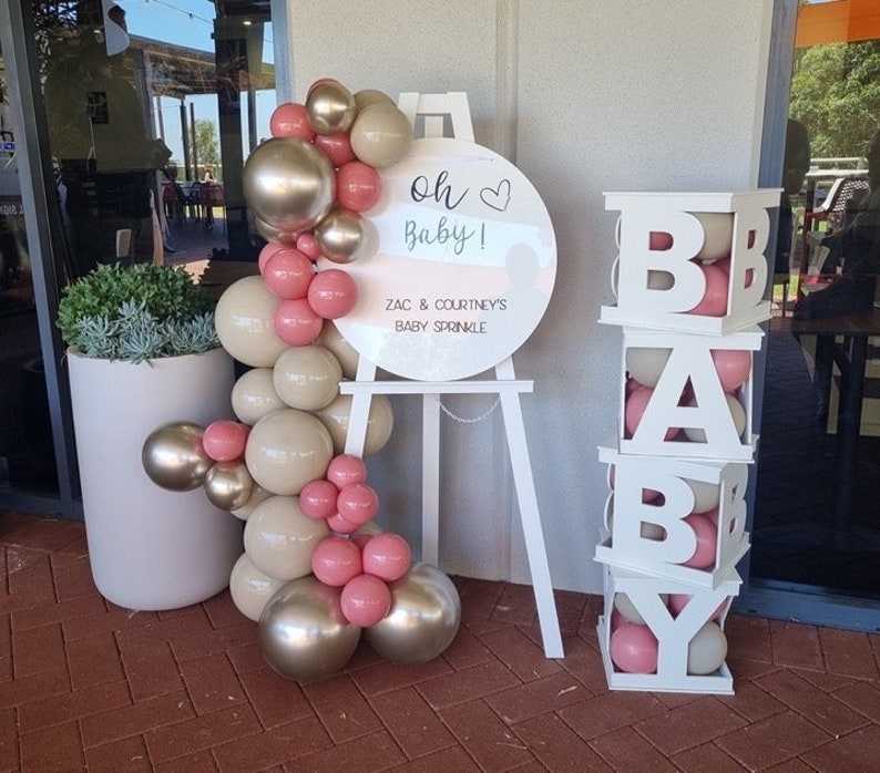 May include: White wooden sign with the text "Oh Baby!" and "Zac & Courtney's Baby Sprinkle".  A white easel holds the sign.  A balloon arch made of pink and beige balloons is on the left.  A white wooden block tower with the letters "BABY" is on the right.