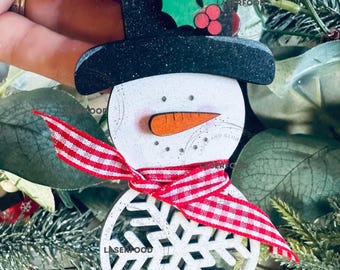 May include: White wooden snowman ornament with a black top hat, red and white striped scarf, and a white snowflake. The hat has a green holly sprig with red berries. The ornament has a red and white striped ribbon with a 2024 tag.