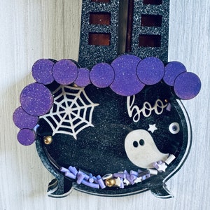 May include: Black cauldron with a purple glitter rim, a white spiderweb, a ghost, and the word "boo" in white. The cauldron is filled with purple and white sprinkles and gold beads.
