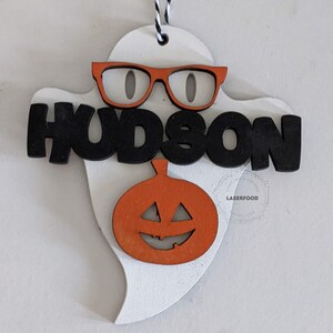 May include: White ghost-shaped wooden ornament with a black "HUDSON" name, orange glasses, and an orange pumpkin with a carved jack-o'-lantern face.