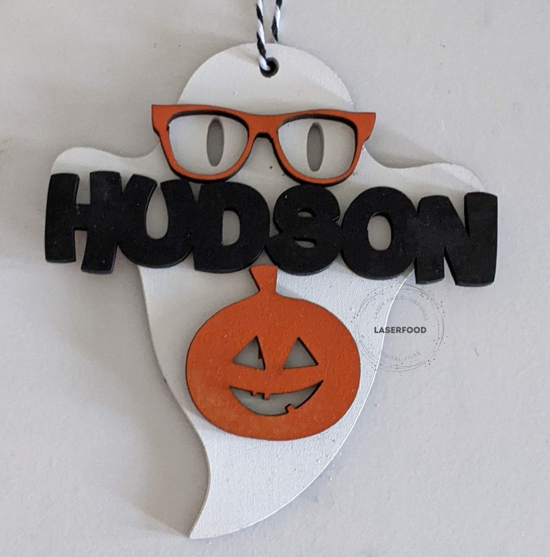 May include: White ghost-shaped wooden ornament with a black "HUDSON" name, orange glasses, and an orange pumpkin with a carved jack-o'-lantern face.