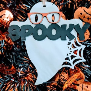 May include: White ghost-shaped ornament with the word "SPOOKY" in black letters and orange glasses. The ornament is surrounded by black and orange tinsel.