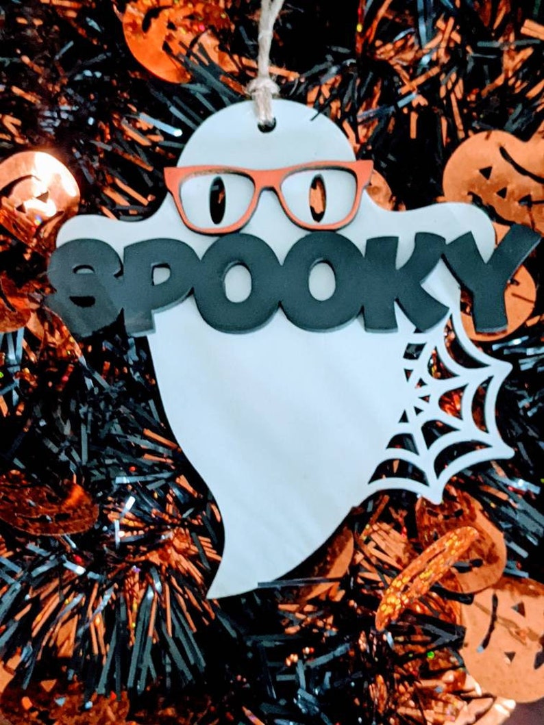 May include: White ghost-shaped ornament with the word "SPOOKY" in black letters and orange glasses. The ornament is surrounded by black and orange tinsel.