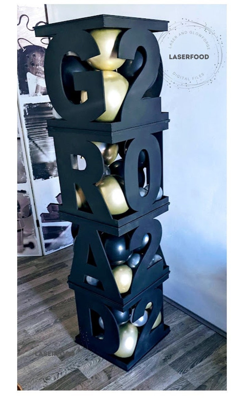 May include: A black wooden box with the letters "GRAD" and the numbers "22" cut out. The box is filled with gold and black balloons. The box is stacked on top of another box with the number "2" cut out. The box is on a wooden floor.