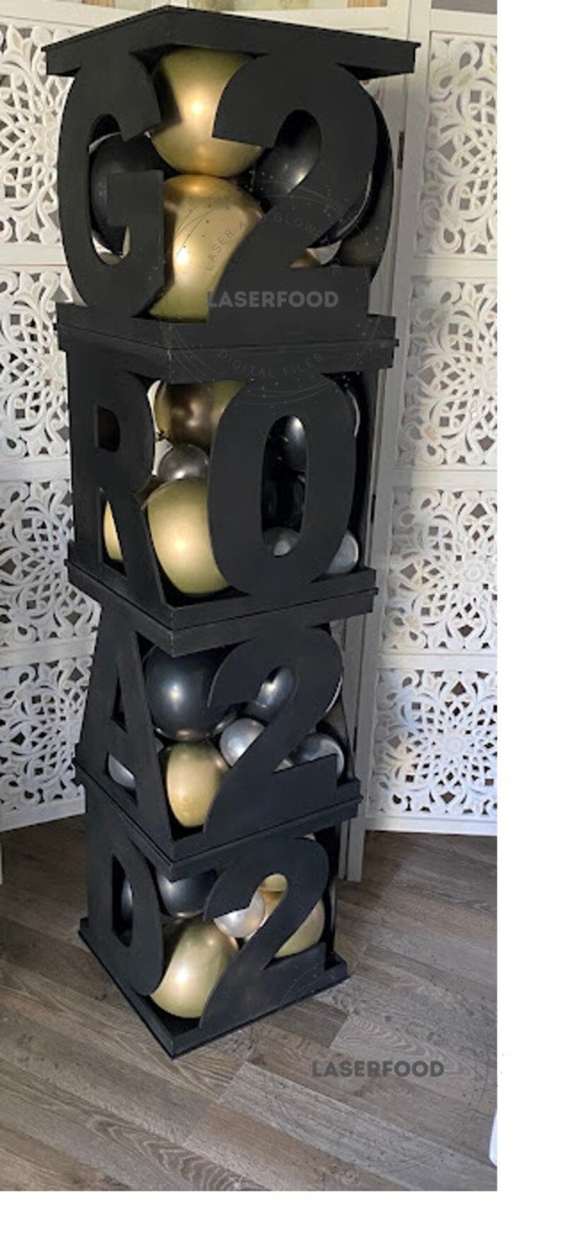 May include: Black wooden boxes stacked to form the number 2022. Each box is filled with gold and silver balloons. The boxes are on a wood floor. The text "LASERFOOD" is printed on the boxes.
