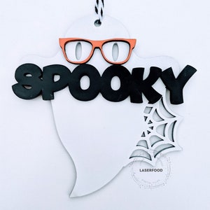 May include: White ghost shaped wooden cutout with the word "spooky" in black letters and orange glasses. A spiderweb design is cut out in the bottom right corner of the ghost.