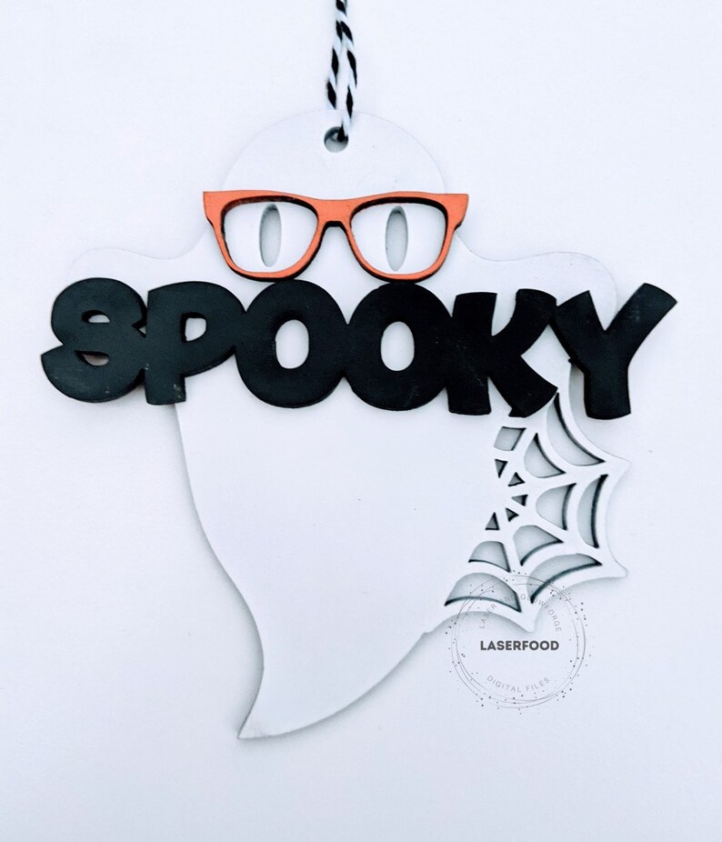May include: White ghost shaped wooden cutout with the word "spooky" in black letters and orange glasses. A spiderweb design is cut out in the bottom right corner of the ghost.