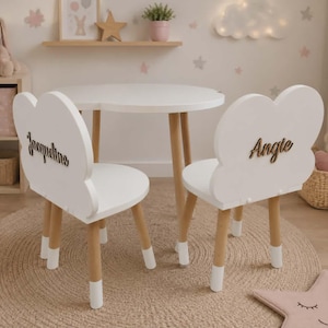 May include: A white children's table and two cloud-shaped chairs. The chairs have wooden legs and backs with the names "Jacqueline" and "Angie" in black script. The table has a white top and wooden legs. The set is on a round, beige rug.